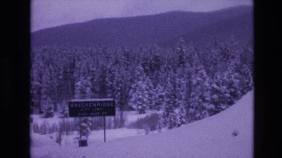 Purple-shifted 8mm frame of a Breckenridge city limit sign beside deep snow, evergreen trees, and a low mountain ridge in Colorado.