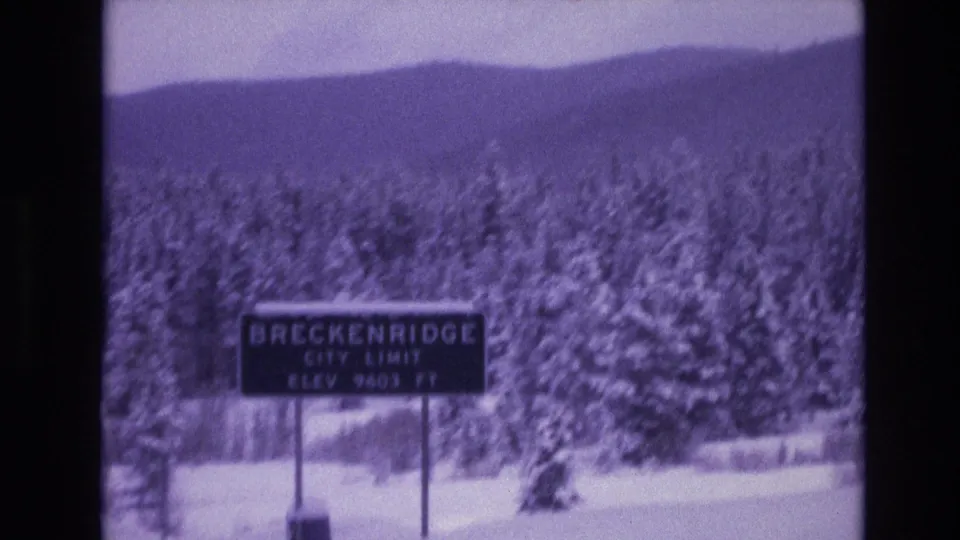 Close roadside view of the Breckenridge city limit sign reading 9603 ft, backed by snow-covered evergreens and a dark mountain ridge in 1970s 8mm footage.