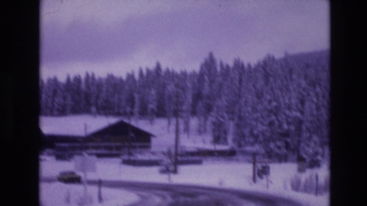 Rustic roadside building near a snow-covered curve in the supplied Vail, Colorado 1974 home-movie footage, with evergreen forest and cloudy winter sky behind it.