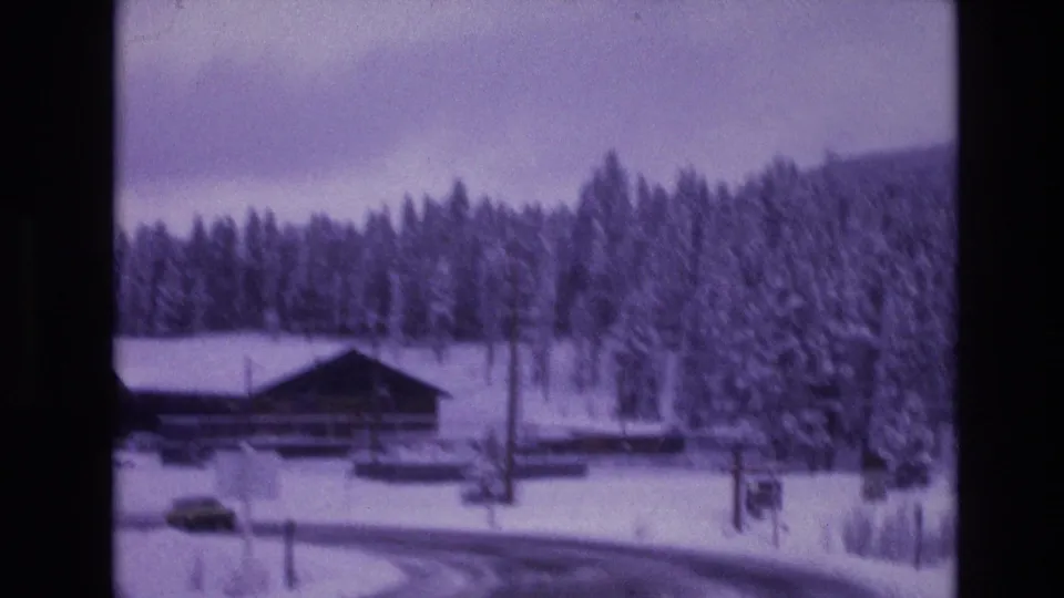 Rustic roadside building near a snow-covered curve in the supplied Vail, Colorado 1974 home-movie footage, with evergreen forest and cloudy winter sky behind it.
