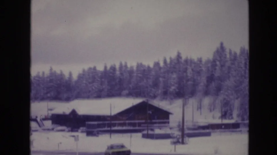 Snow-covered roadside scene from the supplied 1974 Vail, Colorado metadata, showing a car, a rustic building, fences, and dense evergreens in soft 8mm color.
