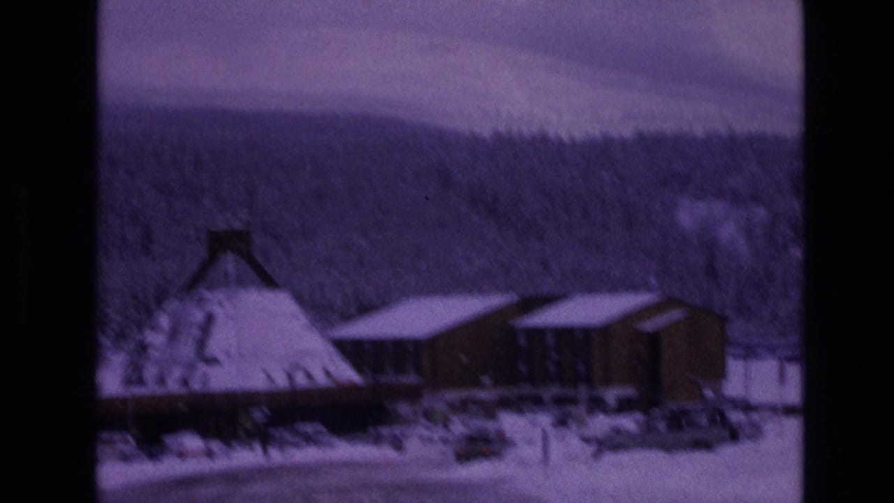 1974 Vail, Colorado 8mm frame showing a snow-covered conical-roof building, adjacent wooden structures, parked cars, evergreen forest, and distant mountains.