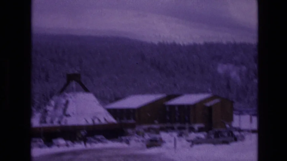 1974 Vail, Colorado 8mm frame showing a snow-covered conical-roof building, adjacent wooden structures, parked cars, evergreen forest, and distant mountains.
