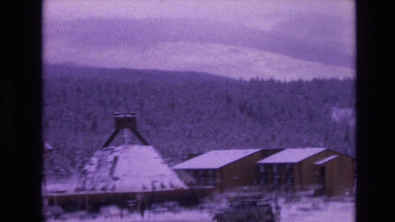 1974 Vail, Colorado home-movie frame with a conical-roof structure, modern wooden building, parked vehicles, snow-covered ground, dense evergreens, and cloudy mountain sky.