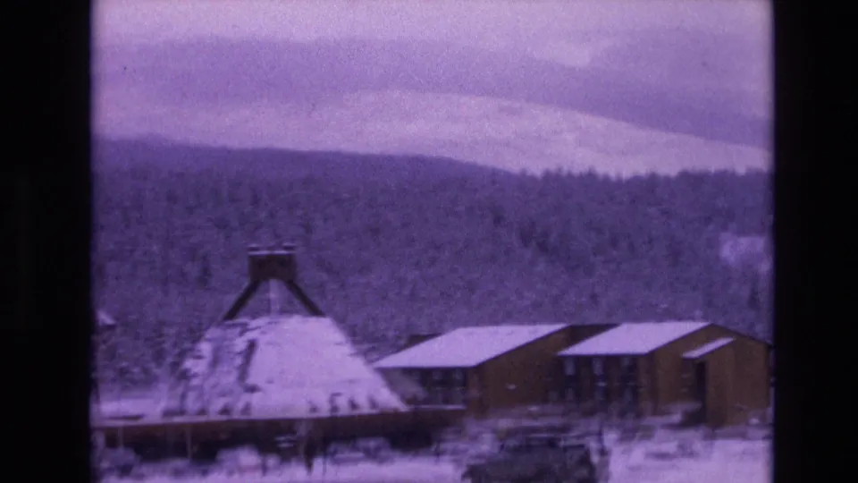1974 Vail, Colorado home-movie frame with a conical-roof structure, modern wooden building, parked vehicles, snow-covered ground, dense evergreens, and cloudy mountain sky.
