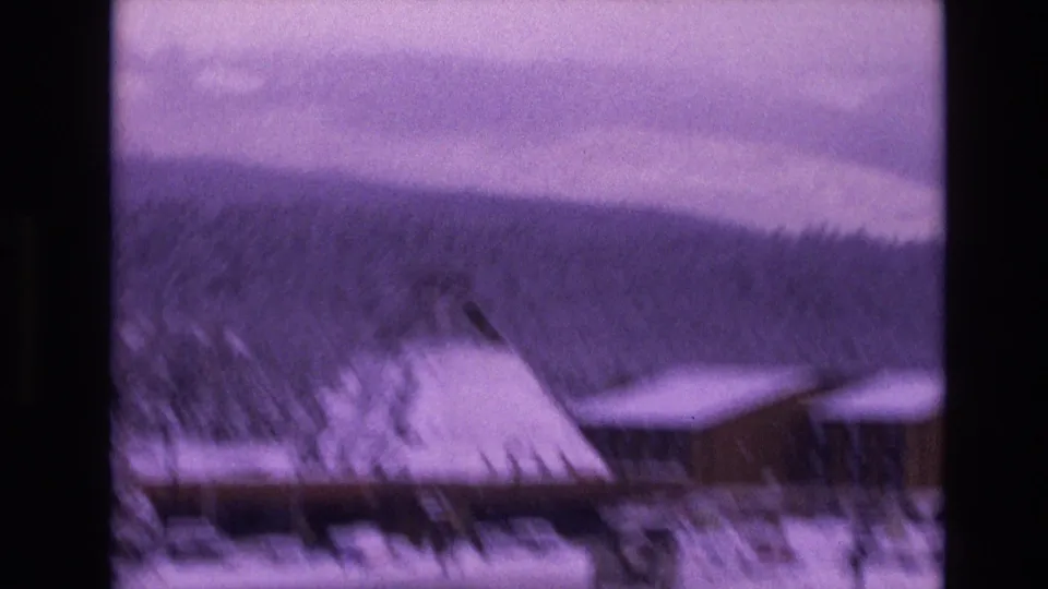 Soft 8mm frame from Vail, Colorado in 1974 showing snowy resort roofs, dark evergreen trees, distant mountain terrain, and visible motion blur from handheld filming.
