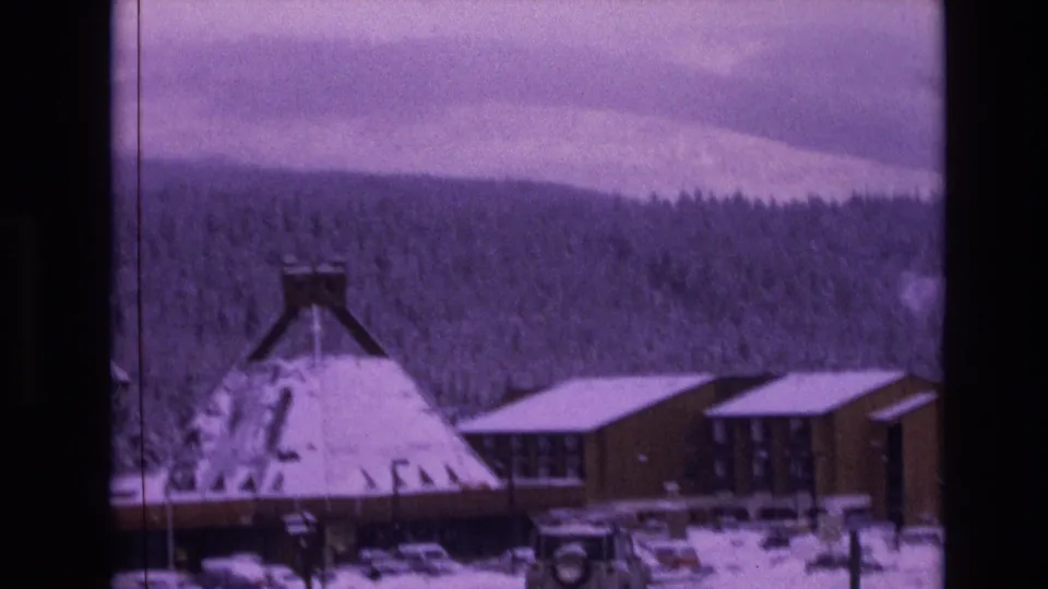 1974 Vail, Colorado 8mm frame showing a snow-covered conical-roof building, adjacent wooden resort structures, several parked cars, evergreen forest, and a pale mountain slope under clouds.