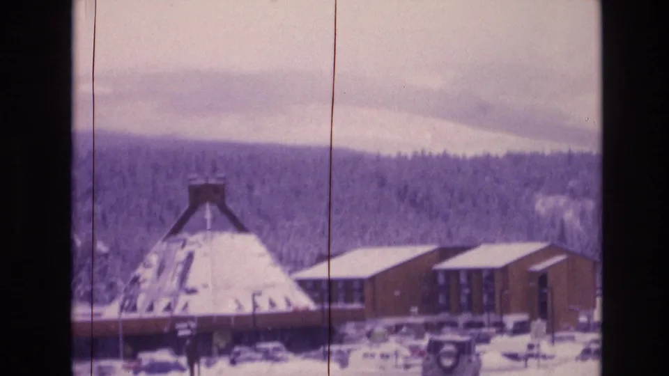 1974 Vail, Colorado 8mm frame with a conical-roof building, adjacent wooden buildings, parked vehicles, snow cover, and distant forested hills.