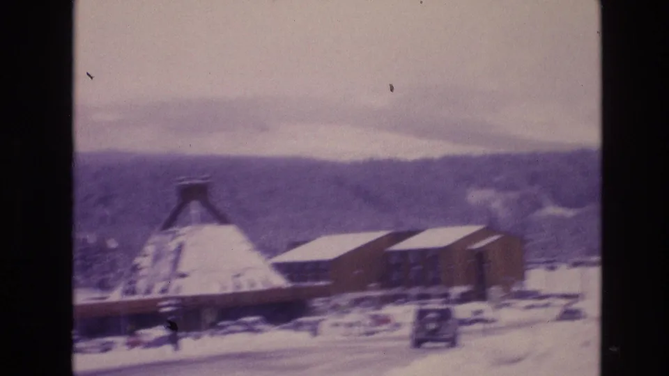 1974 Vail, Colorado 8mm frame showing a wider snowy roadside view with mountain buildings, a parked vehicle, and wooded hills under a cloudy sky.