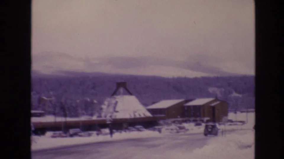1974 Vail, Colorado 8mm frame with snow-covered roadside buildings, a parked vehicle, wooded hills, and a small distant person walking through the scene.