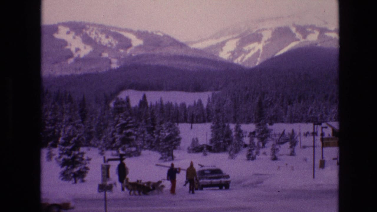 1974 Vail, Colorado 8mm frame showing bundled people and dogs near a parked car in a snowy roadside area below forested slopes and distant ski runs.