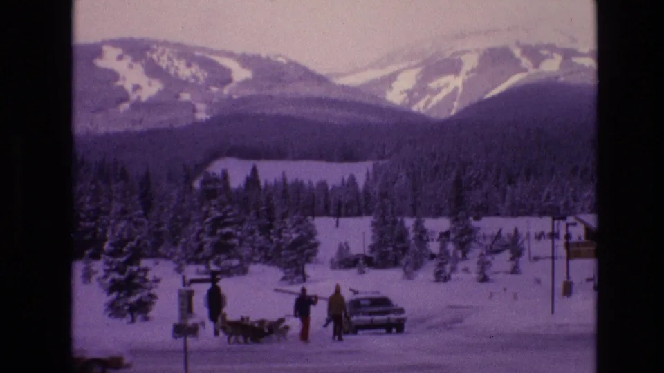 1974 Vail, Colorado 8mm frame showing bundled people and dogs near a parked car in a snowy roadside area below forested slopes and distant ski runs.
