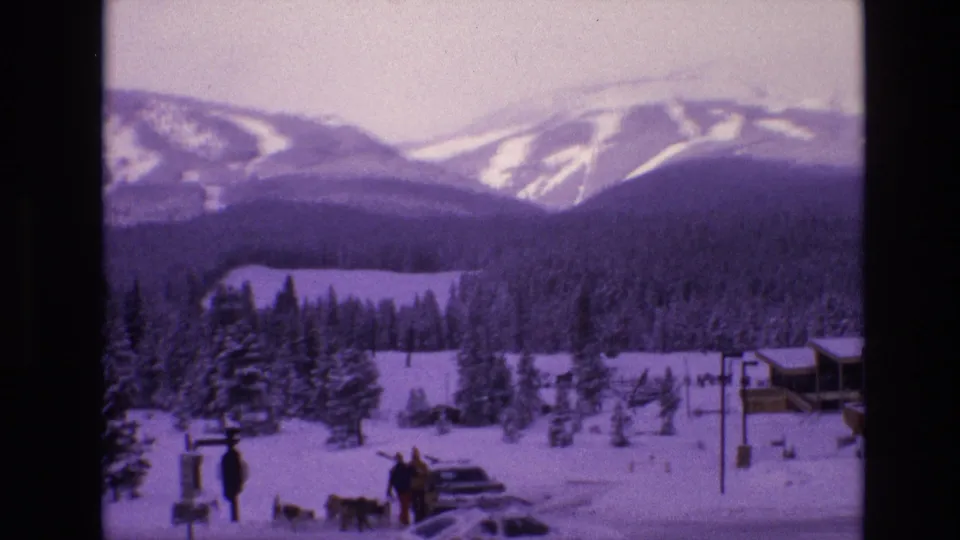 1974 Vail, Colorado home-movie frame with people and dogs gathered near a parked car, snow-covered evergreens, low buildings, and ski-marked mountain slopes beyond.