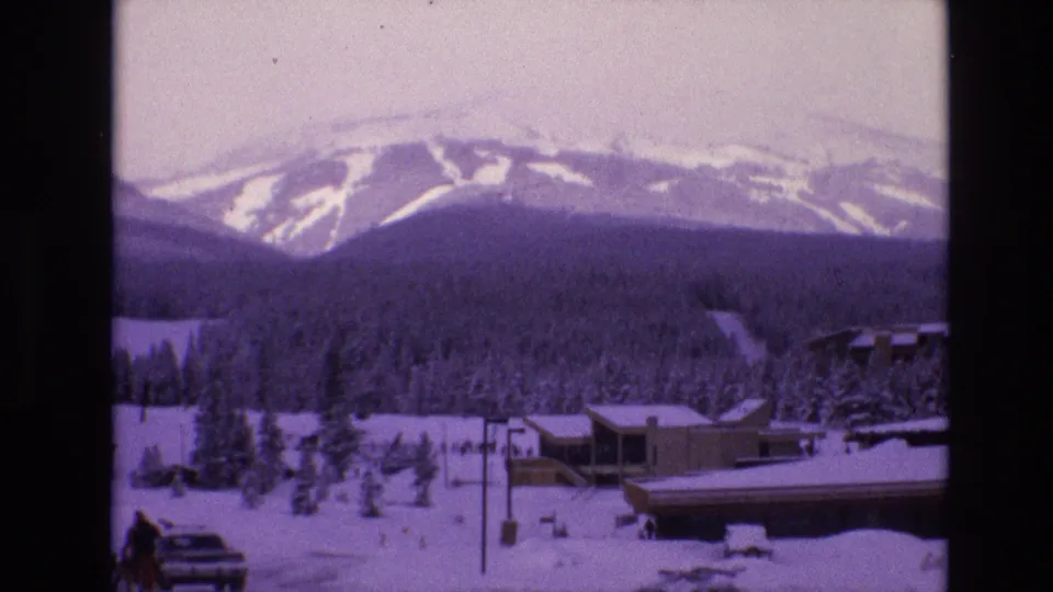 1974 Vail, Colorado 8mm frame looking across snow, evergreens, roadside posts, resort buildings, and distant ski runs, with people and a car partly visible near the bottom edge.