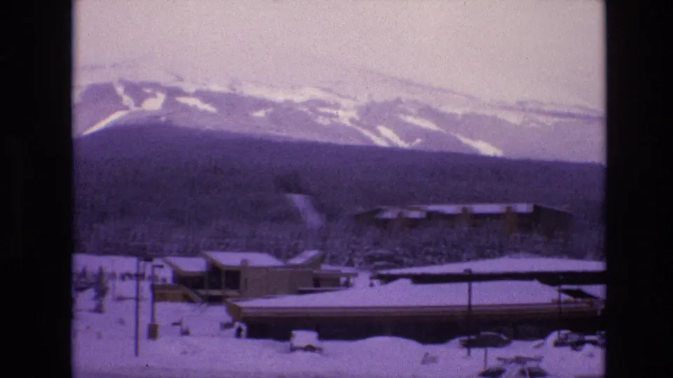 1974 Vail, Colorado 8mm frame showing low snow-covered resort buildings, roadside posts, evergreens, and ski runs on the mountain ridge in the background.