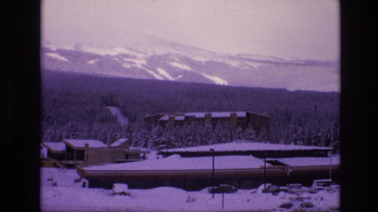 Vail, Colorado winter resort view from 1974 8mm footage, with snowy low buildings, evergreen forest, and mountains under a pale sky.