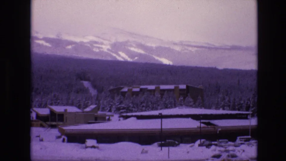Vail, Colorado winter resort view from 1974 8mm footage, with snowy low buildings, evergreen forest, and mountains under a pale sky.