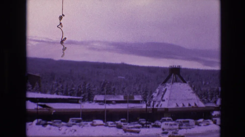 Vail, Colorado 1974 frame with an A-frame resort building, snow-covered foreground cars, evergreen woods, and layered mountains in the distance.