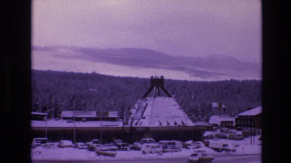 1974 Vail, Colorado resort view showing an A-frame lodge-like structure, snow-covered cars, evergreen forest, and a hazy mountain ridge.