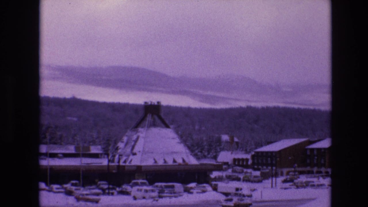 1974 8mm view of Vail, Colorado, showing a conical-roofed snow-covered building, parked cars, evergreen woods, and distant snowy ridges under a pale sky.
