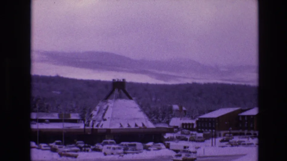 1974 8mm view of Vail, Colorado, showing a conical-roofed snow-covered building, parked cars, evergreen woods, and distant snowy ridges under a pale sky.