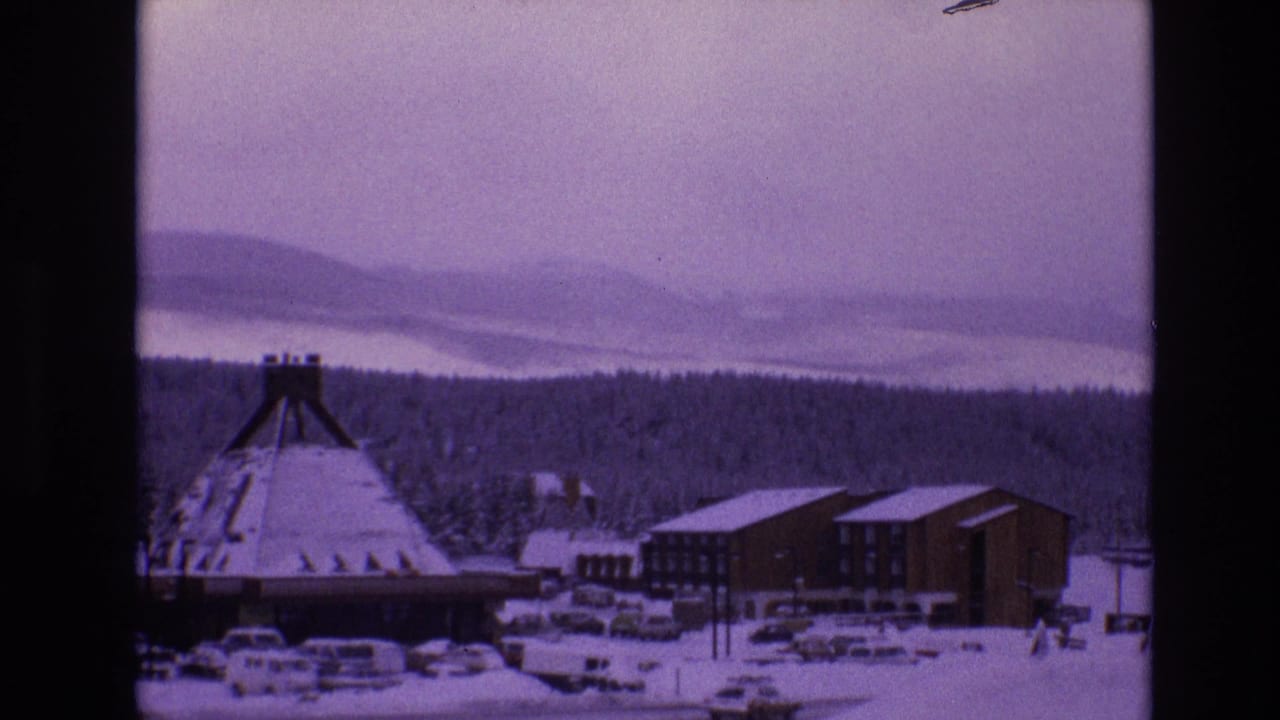 Snowy conical-roofed building beside lodges and parked cars.