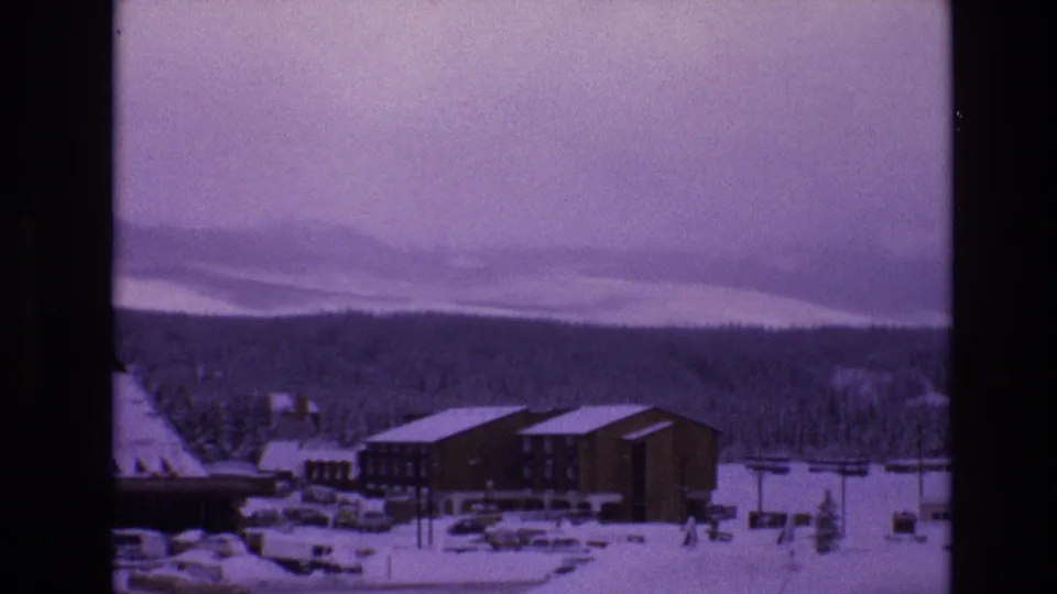 1974 Vail 8mm frame showing lodge-style buildings, snow-covered lots, parked cars, evergreen trees, and misty mountain slopes in the distance.