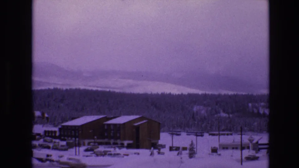Late pan frame from 1974 Vail, Colorado 8mm footage, with lodge buildings, snowy grounds, parked vehicles, utility poles, evergreen woods, and hazy mountain ridges.