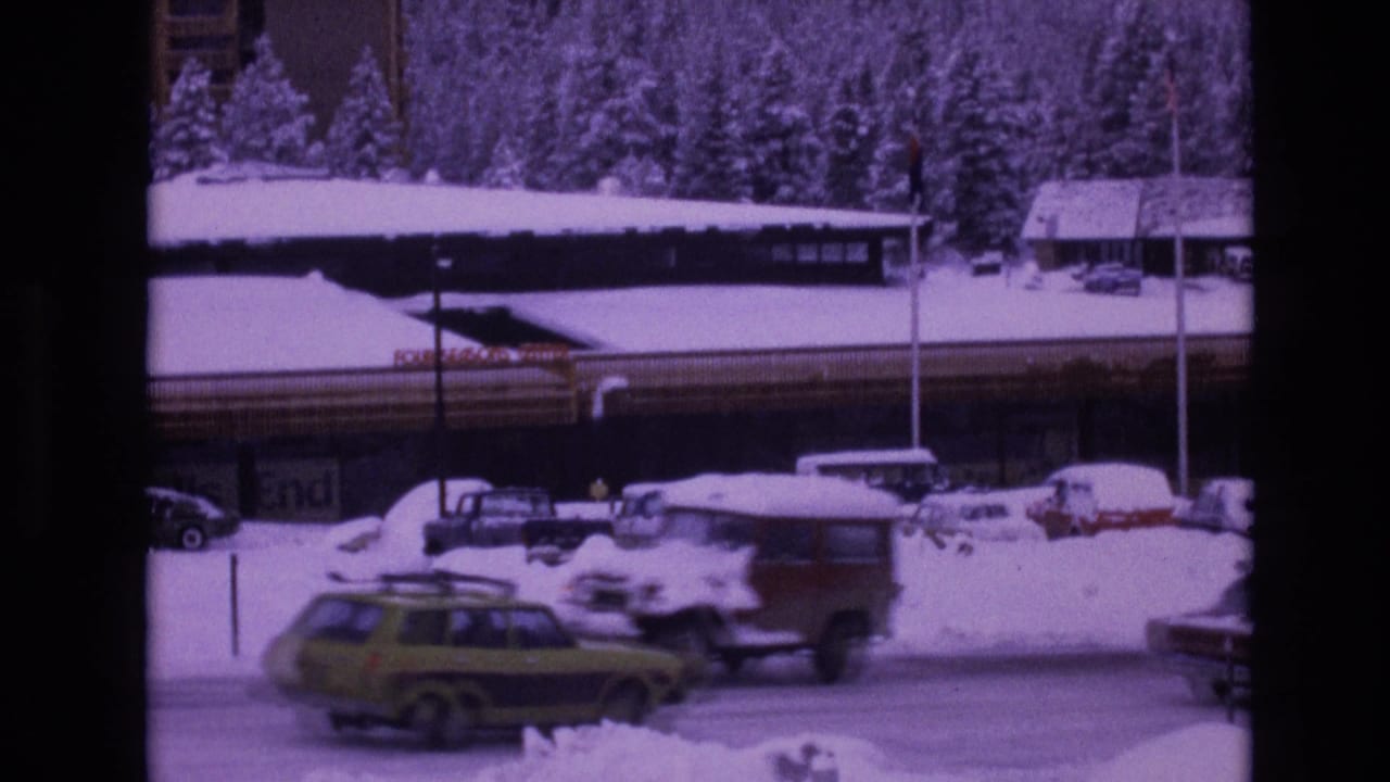 Snowy roadside parking area with cars and a lodge building.