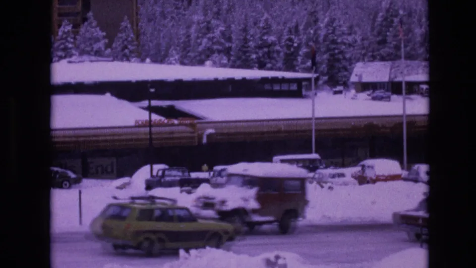 A snowy roadside scene in Vail shows a low lodge-like building, parked 1970s cars, a station wagon, snowbanks, evergreen trees, and a partially visible sign.