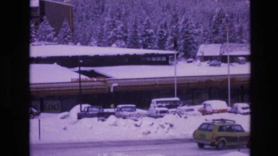The frame shows a snow-packed Vail roadside with several parked cars, a long dark-roofed building, evergreen trees, a pole, and a green station wagon at the edge of the road.