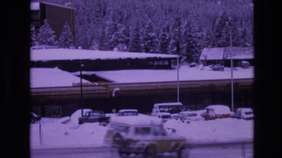A moving vehicle blurs past a snowy Vail roadside where parked cars, snowbanks, a low resort building, evergreen trees, and a tall pole remain in view.