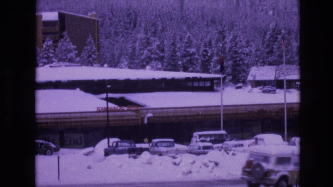 1974 Vail, Colorado home-movie frame showing a snow-covered road edge, parked vintage cars, a long resort-style building with signage, and dense evergreen trees in winter.