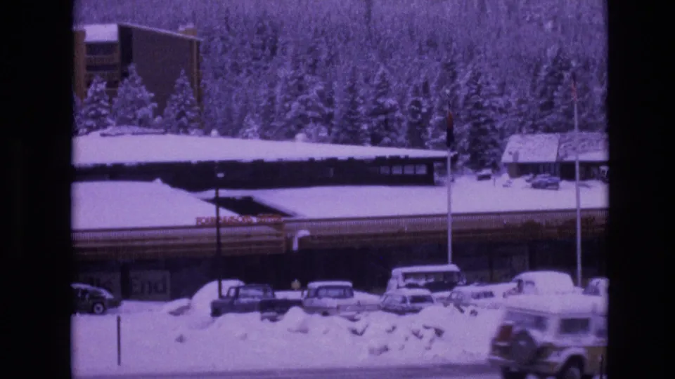 1974 Vail, Colorado home-movie frame showing a snow-covered road edge, parked vintage cars, a long resort-style building with signage, and dense evergreen trees in winter.