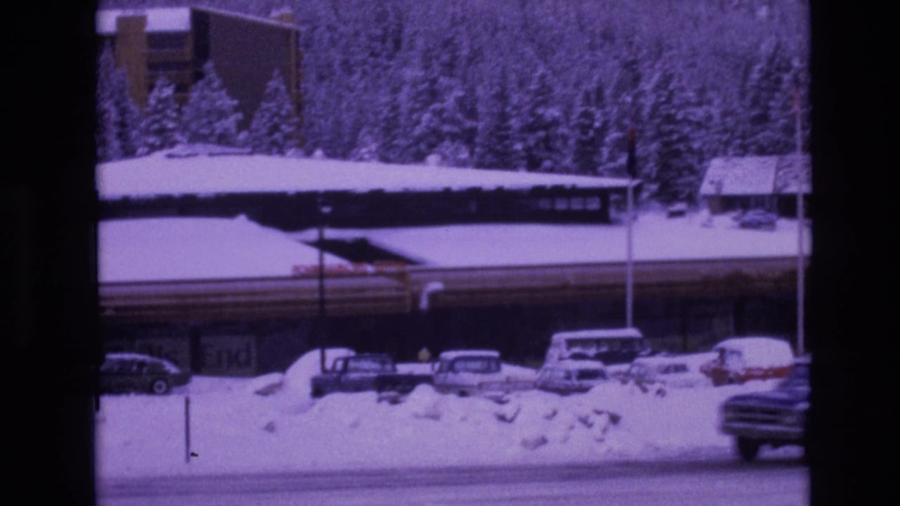 A 1974 Vail, Colorado winter frame with snowbanks, a road, parked cars, a long building with visible signage, and a wooded slope behind the resort area.