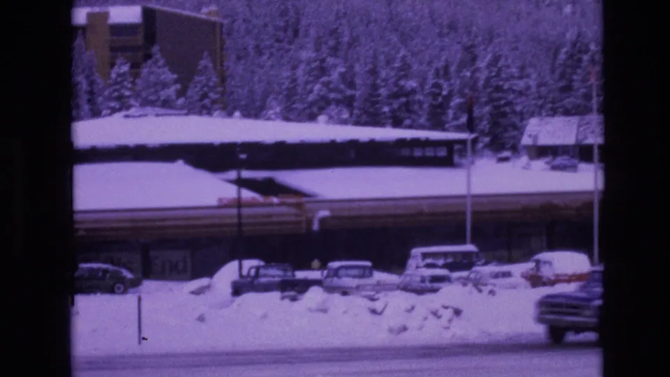 A 1974 Vail, Colorado winter frame with snowbanks, a road, parked cars, a long building with visible signage, and a wooded slope behind the resort area.