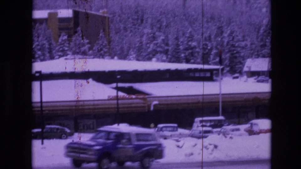 1974 Vail, Colorado frame showing a blue vintage vehicle moving across the foreground, with snowbanks, parked cars, a long building, and evergreen trees behind it.