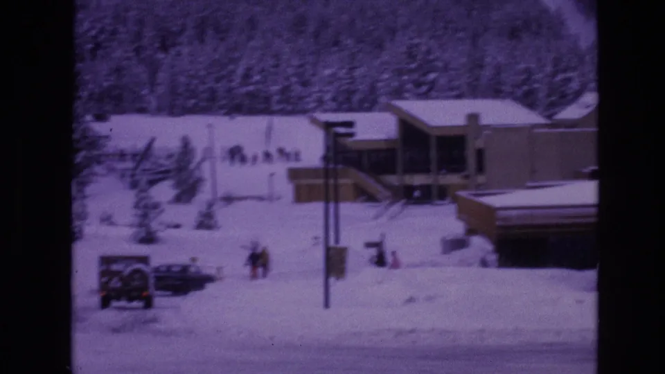 A 1974 Vail, Colorado winter frame with modern resort buildings, parked vehicles, heavy snow cover, poles, and distant people walking through the snowy area.