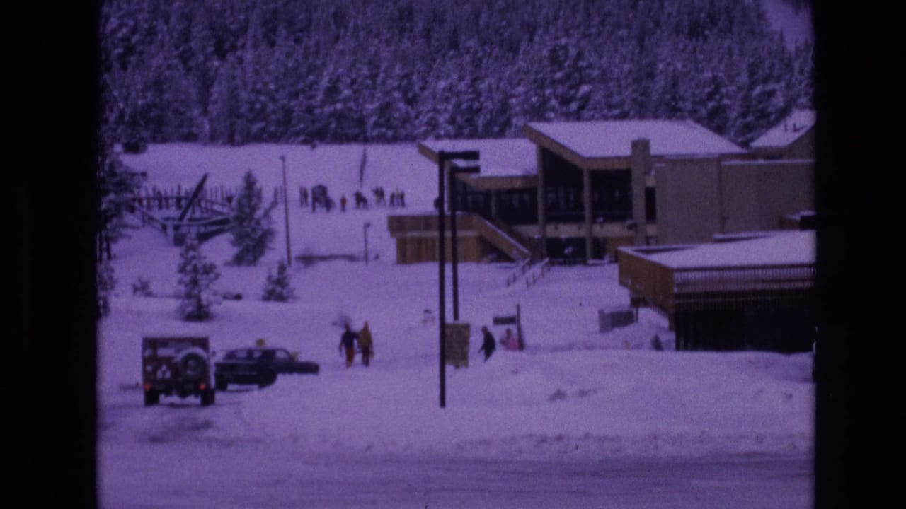 1974 Vail, Colorado home-movie frame showing a snowy roadside with parked cars, a few bundled pedestrians, utility poles, small evergreens, and a larger lodge-like building in the background.