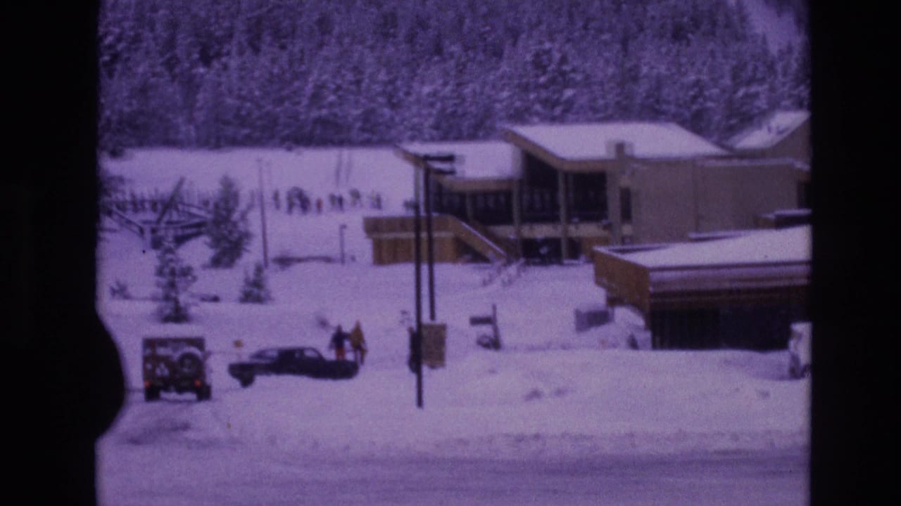 1974 Vail, Colorado frame with a snow-covered street, parked cars, small groups of pedestrians in winter clothing, utility poles, evergreens, and a lodge-style structure under overcast light.