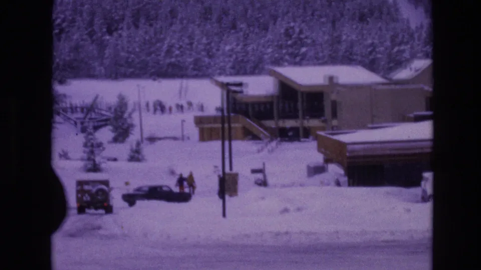 1974 Vail, Colorado frame with a snow-covered street, parked cars, small groups of pedestrians in winter clothing, utility poles, evergreens, and a lodge-style structure under overcast light.