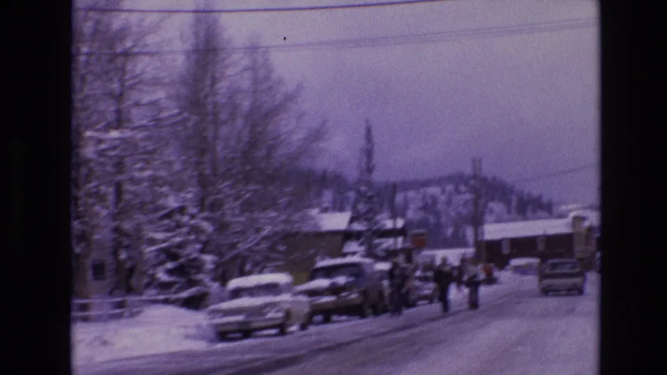1974 Vail, Colorado home-movie view down a snowy street with parked cars, bundled pedestrians, snow-laden trees, low buildings, utility wires, and hazy mountain slopes in the distance.