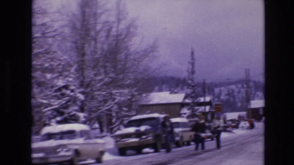 1974 Vail, Colorado frame showing a snow-covered town street with parked cars along the roadside, bundled pedestrians walking nearby, leafless trees, low buildings, and mountain terrain beyond.