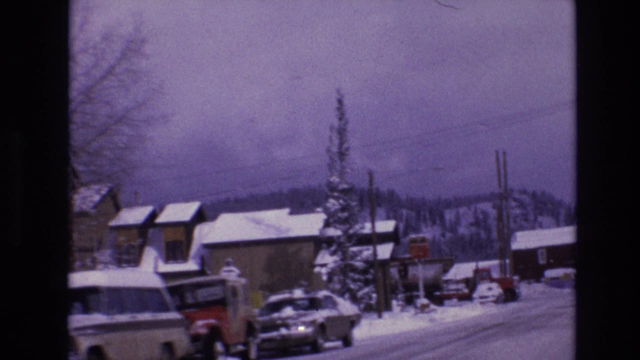 Snowy roadside with pickup trucks, cars, buildings, utility poles, and distant hills.