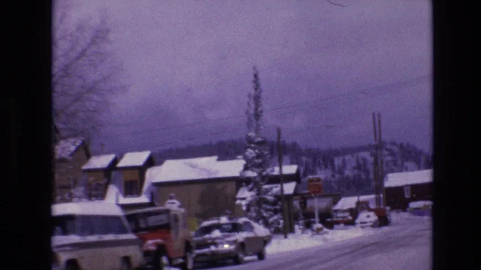 1974 Vail, Colorado roadside scene with parked pickup trucks and cars in front of snow-covered buildings, utility poles, and distant winter hills.