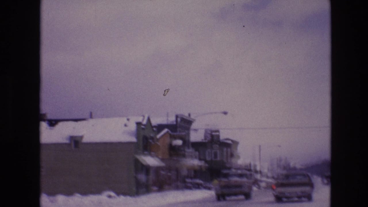 Cars travel along a snowy Vail business district street in 1974, with storefront buildings, streetlights, and a muted winter sky visible through the 8mm frame.