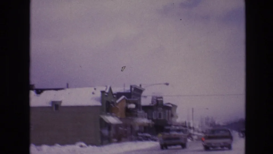 Cars travel along a snowy Vail business district street in 1974, with storefront buildings, streetlights, and a muted winter sky visible through the 8mm frame.