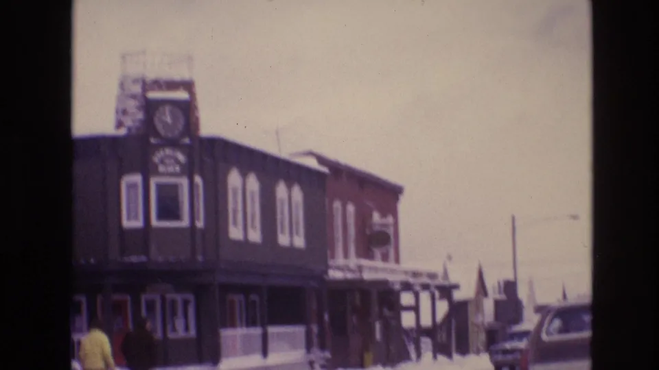 A prominent clock-tower building in Vail's snowy 1974 business district appears beside pedestrians, parked cars, storefront windows, and winter street fixtures.