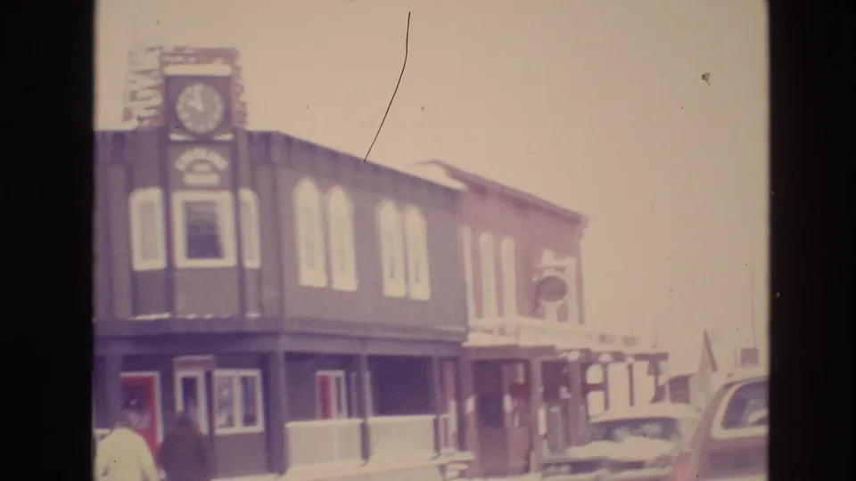 The later frame holds on the Vail clock-tower storefront and adjacent snowy sidewalk, with pedestrians, cars, rooflines, and bright 8mm winter exposure.