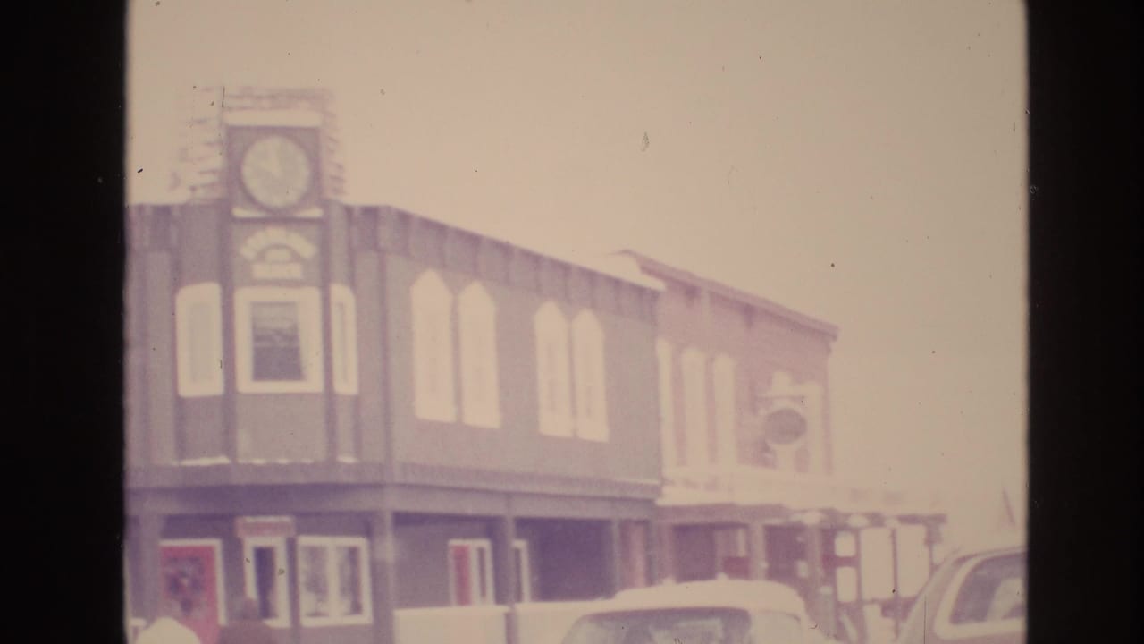 Faded 8mm frame from Vail, Colorado showing a two-story street building with a clock-like facade detail, arched upper windows, and parked cars in the foreground.